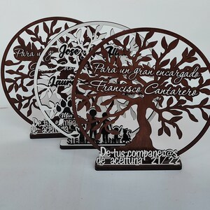 以下が含まれることがあります： 白い文字で書かれた木製のツリー型のプレートが3つ。プレートには、枝と葉のある木と、「Para un gran encargado Francisco Cantarero」と「De tus companeras de aceituna 21/22」の文字が書かれています。