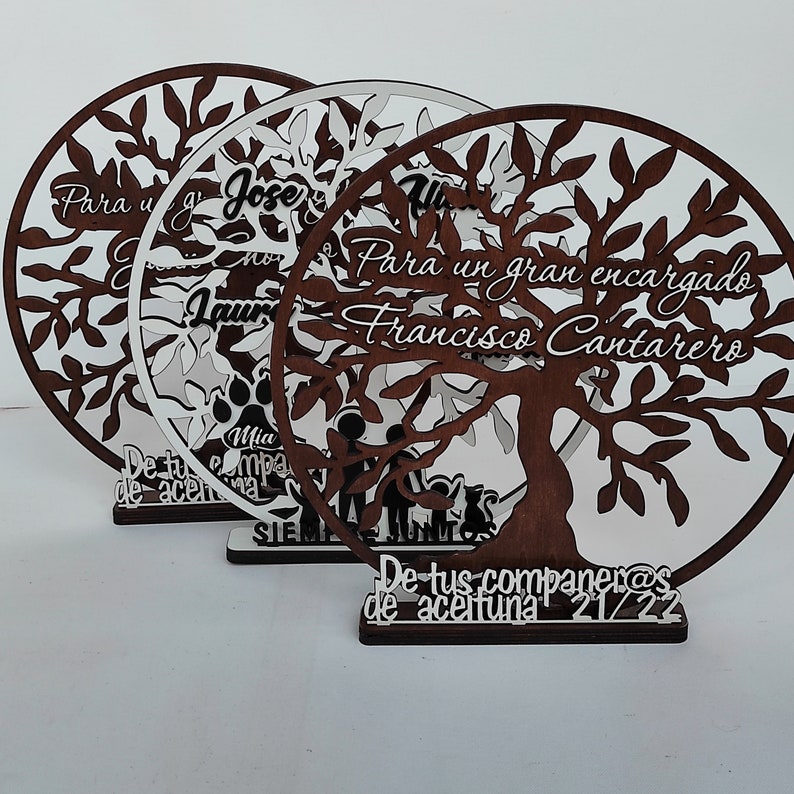 以下が含まれることがあります： 白い文字で書かれた木製のツリー型のプレートが3つ。プレートには、枝と葉のある木と、「Para un gran encargado Francisco Cantarero」と「De tus companeras de aceituna 21/22」の文字が書かれています。