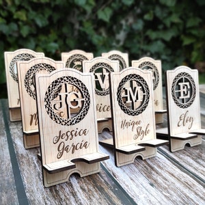 Op de afbeelding: Een set van acht houten telefoonstandaards met lasergesneden ontwerpen. Elke standaard heeft een uniek monogram en een gepersonaliseerde naam, zoals "Jessica Garcia", "Unique Style" en "Eloy".