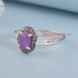 May include: A silver ring with a pink oval gemstone surrounded by a halo of smaller diamonds. The ring is on a light blue background.