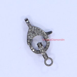 Könnte beinhalten: Ein silberfarbener Verschluss mit einer runden Öffnung und einer kleinen Schlaufe an der Seite. Der Verschluss ist mit kleinen, klaren Diamanten besetzt.