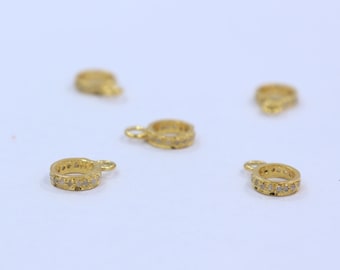 May include: Several small, gold-colored ring pendants with a loop at the top. Each ring is encrusted with small, clear stones. The pendants are scattered on a white surface, with some in focus and others blurred.