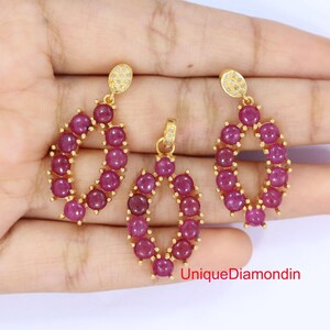 May include: A gold-toned jewelry set featuring a pendant and a pair of earrings. Each piece is adorned with oval-shaped clusters of deep red gemstones. The earrings and pendant have a small, round, diamond-studded top. The text "UniqueDiamondin" is visible.