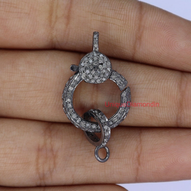 May include: A silver-toned clasp with a round, pave-set diamond design. The clasp is made of metal and has a loop on one side and a hook on the other.