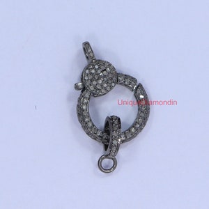 May include: A silver-toned clasp with a round design, featuring small, clear diamonds set in the metal. The clasp has a spring-loaded mechanism and a small loop for attaching to a chain or cord.