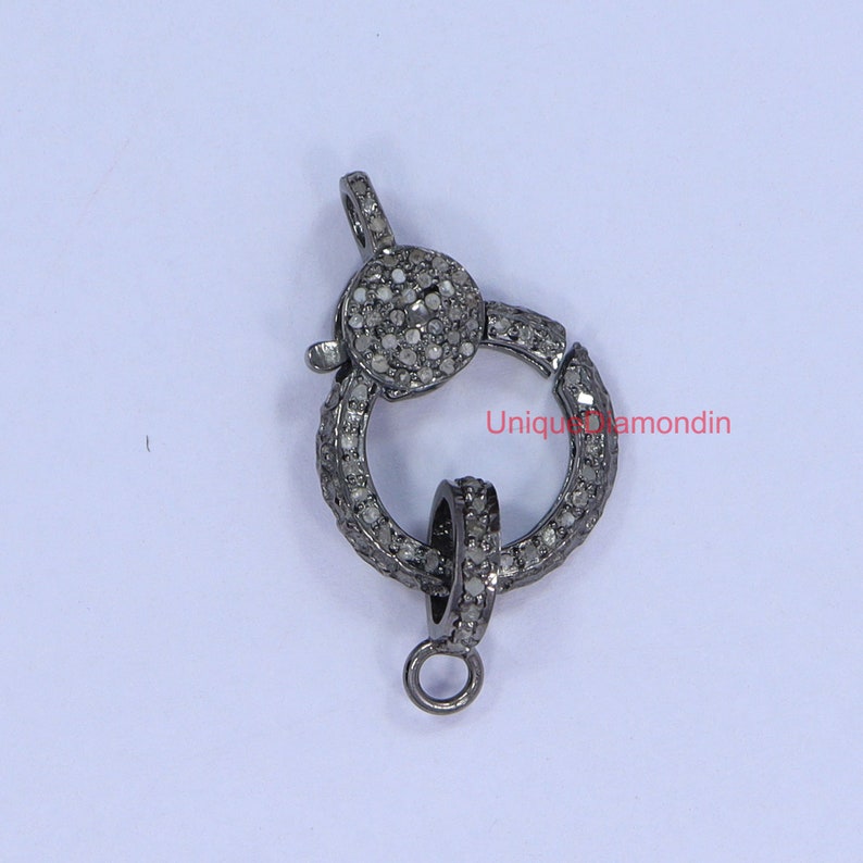 May include: A silver-toned clasp with a round design, featuring small, clear diamonds set in the metal. The clasp has a spring-loaded mechanism and a small loop for attaching to a chain or cord.