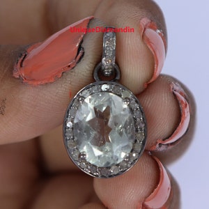 May include: A silver pendant with a large oval-shaped clear gemstone surrounded by smaller diamonds. The pendant is held in a hand with painted nails.