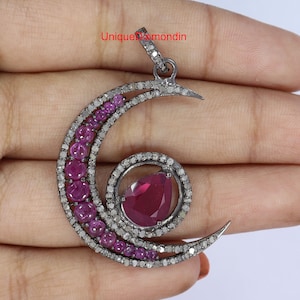 May include: A crescent moon pendant with a large, pear-shaped ruby at the center. The pendant is adorned with smaller, round rubies and diamonds, set in a dark metal. The text "UniqueDiamondin" is visible.