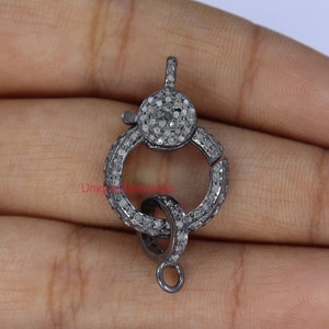 May include: A silver-toned clasp with a round, pave-set diamond design. The clasp has a small loop at the bottom for attaching to a chain or cord.