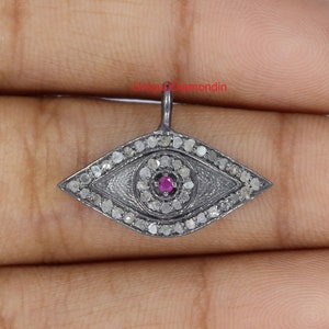 Puede incluir: Pendiente de plata en forma de ojo turco con una piedra preciosa roja en el centro y diamantes engastados en pavé alrededor del ojo. El pendiente está sobre un fondo blanco.