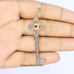 May include: A silver key pendant featuring a red gemstone at its centre, surrounded by small diamonds. The key is set in a gold tone and has an ornate design.