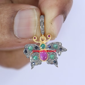 May include: A gold butterfly pendant with a pink ruby gemstone in the center, surrounded by green emeralds and blue sapphires. The pendant is set with small diamonds and has a loop for attaching a chain.