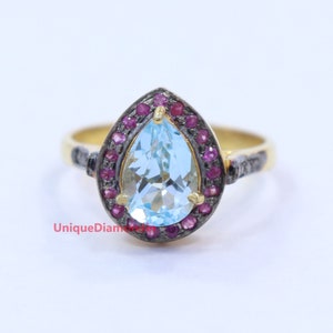 Może przedstawiać: Złoty pierścionek z centralnym, jasnoniebieskim kamieniem szlachetnym w kształcie gruszki, otoczonym małymi, czerwonymi kamieniami. Obrączka pierścionka ma ciemnoszary akcent. Widoczny jest tekst "UniqueDiamonds".