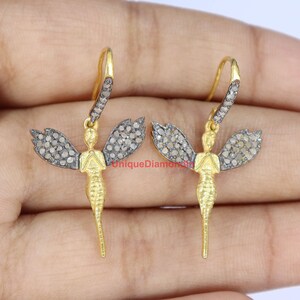 May include: A pair of gold dangle earrings featuring a fairy design with wings set with small diamonds. The fairy is standing with hands clasped in front of her.