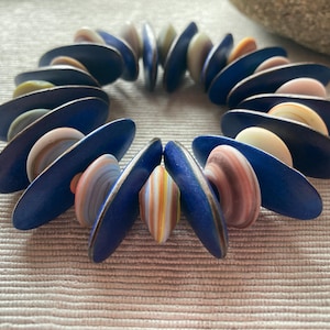 May include: A beaded bracelet featuring navy blue, oval-shaped beads interspersed with colorful, striped beads. The bracelet is made of various colors, including blue, white, pink, and orange. The bracelet is displayed on a textured, neutral-colored surface.