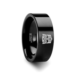 Könnte beinhalten: Schwarzer Ring aus Wolframcarbid mit einem silbernen, gravierten Transformers-Logo. Der Ring ist mit "TUNGSTEN CARBIDE" beschriftet.