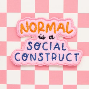 Könnte beinhalten: Rosa Filz-Patch mit dem Text "NORMAL is a SOCIAL CONSTRUCT" in orange, blau und weißer Stickerei.