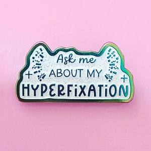 Op de afbeelding: Een witte emaille pin met een regenboogkleurige, iriserende afwerking. De pin heeft de tekst "Ask me about my hyperfixation" in zwarte letters.