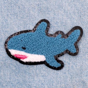 Op de afbeelding: Een blauwe en zwarte vilt haai patch met een witte mond en roze tanden. De patch is perfect om een ​​leuke en fantasierijke touch toe te voegen aan kleding, tassen of andere accessoires.