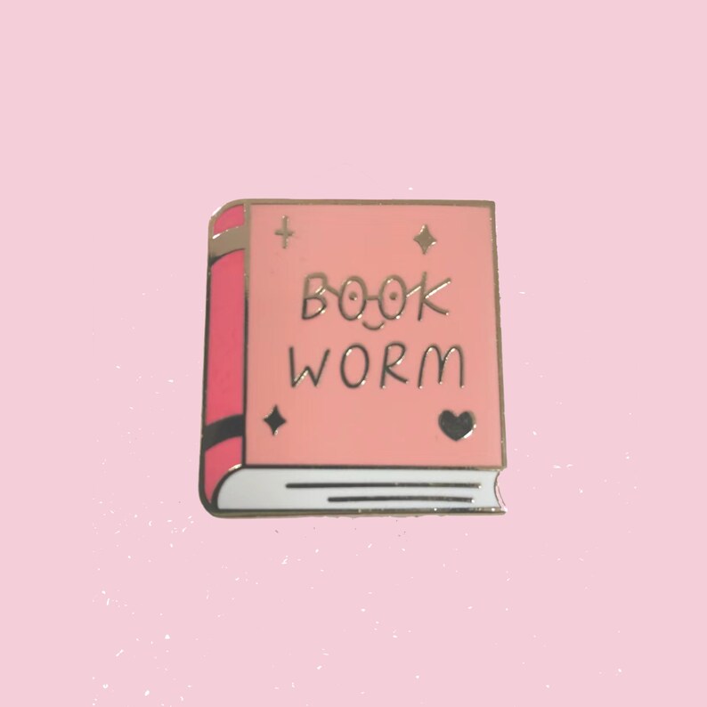 May include: Pink enamel pin shaped like a book with the text "BOOK WORM" in gold lettering. The book has a gold heart and stars on the cover.