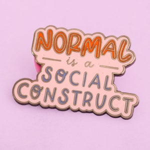 May include: A pink enamel pin with the text "NORMAL is a SOCIAL CONSTRUCT" in orange and blue lettering.