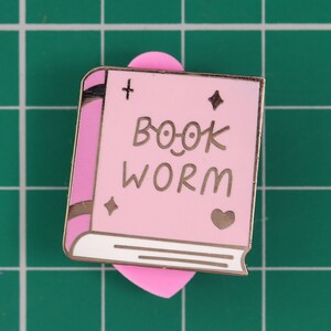 May include: Pink enamel pin shaped like a book with the text "BOOK WORM" in gold lettering. The book has a gold bookmark in the shape of a heart.