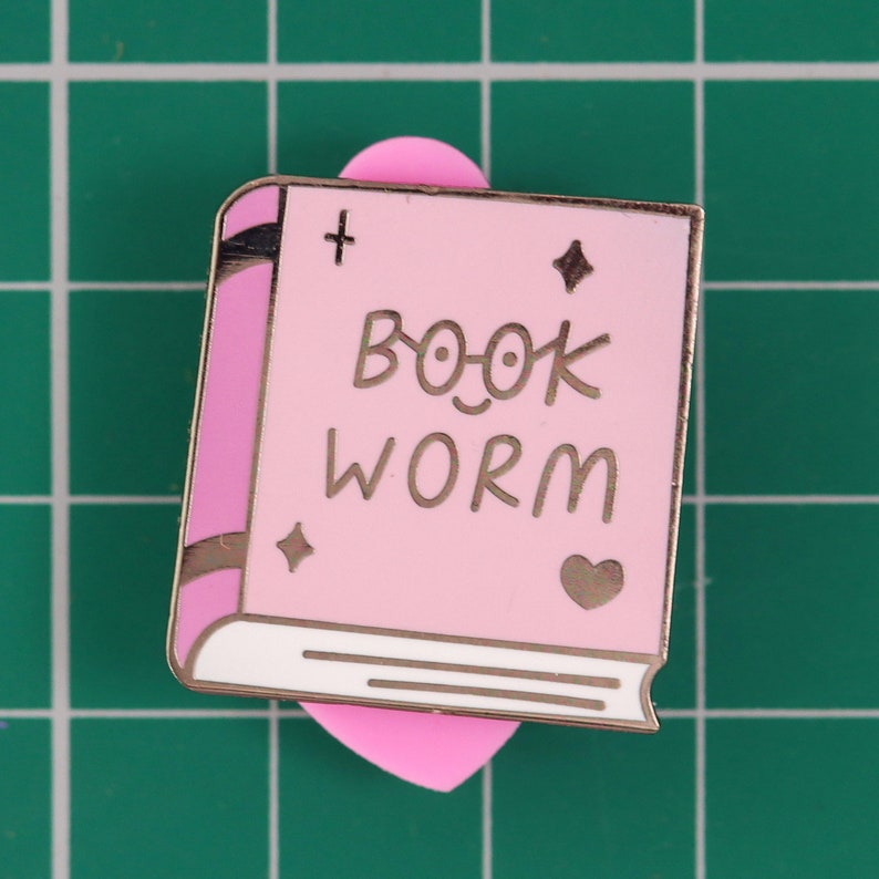 May include: Pink enamel pin shaped like a book with the text "BOOK WORM" in gold lettering. The book has a gold bookmark in the shape of a heart.