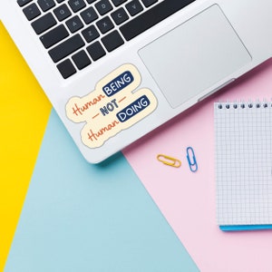 Może przedstawiać: Biały laptop z dwoma naklejkami w prawym g&oacute;rnym rogu. Naklejki są białe z niebieskim i czerwonym tekstem. Tekst na naklejkach brzmi "Human Being - Not Human Doing". Laptop leży na r&oacute;żowo-niebieskiej powierzchni z białym notesem i dwoma spinaczami.