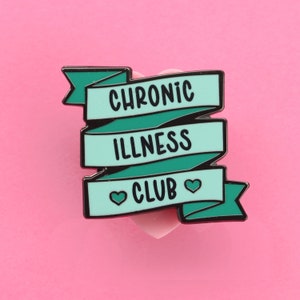May include: A teal blue enamel pin with a black outline. The pin is shaped like a banner with the words "Chronic Illness Club" and two small hearts.