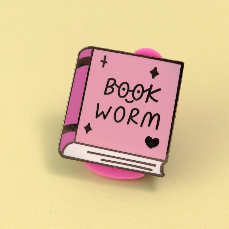 May include: Pink enamel pin shaped like a book with the text "Book Worm" and a heart. The pin has a gold finish and is attached to a pink rubber backing.
