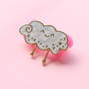 May include: A gold-colored enamel pin shaped like a white cloud with a smiling face and two gold stars. The cloud has a swirl design and small white stars.