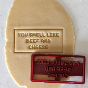 May include: Red plastic cookie cutter with the text "YOU SMELL LIKE BEEF AND CHEESE" printed on it. The cookie cutter is on top of a round piece of cookie dough.
