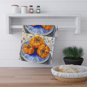May include: A kitchen towel with a watercolour painting of ripe persimmons on blue plates. The persimmons are a vibrant orange colour, and the plates have a blue and white design. The towel is hanging on a white shelf.