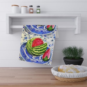 Könnte beinhalten: Ein Küchentuch mit einer Aquarellillustration einer Wassermelone auf einem blau-weißen Teller. Die Wassermelone ist aufgeschnitten und zeigt ihr rotes Fruchtfleisch. Das Tuch hängt an einem weißen Regal.