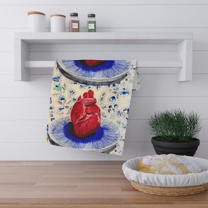 May include: A kitchen towel with a realistic red heart design on a blue plate. The towel has a floral pattern in blue and green on a cream background. The towel is hanging on a white shelf.