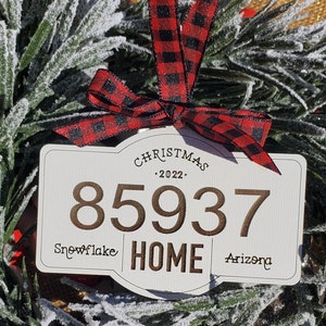 Puede incluir: Adorno navideño blanco con una cinta de cuadros rojos y negros. El adorno tiene el número dorado "85937" y el texto "Snowflake Home Arizona" con el año "2022" sobre el número.