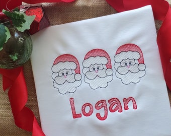 May include: White t-shirt with three embroidered Santa faces and the name "Logan" in red.