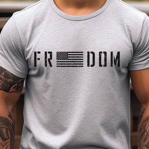 May include: A light grey short-sleeved t-shirt featuring the word FREEDOM printed in a distressed black font across the chest. The letter E is replaced by a graphic of the American flag, also rendered in a distressed black and white style.
