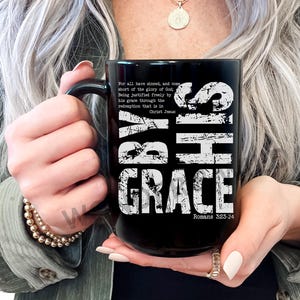 May include: A black coffee mug with the words "BY HIS GRACE" in large white distressed lettering. The mug also features a Bible verse. The mug is held by a person.