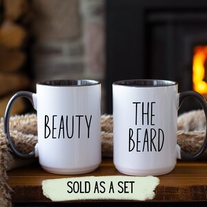 Two white coffee mugs with black handles and black rims. The mug on the left has the word 'BEAUTY' printed on it. The mug on the right has the words 'THE BEARD' printed on it. The mugs are on a wooden surface. The text 'SOLD AS A SET' is printed on a green banner below the mugs.