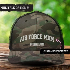 May include: A camouflage trucker hat with black mesh back. The hat has white embroidery that says "Air Force Mom Morrison" with a pink arrow pointing to the text. The text "Custom Embroidery" is also visible on the hat.