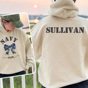 May include: A cream-colored hoodie with the text "NAVY MOM" and a camouflage bow on the front. The back of the hoodie has the text "SULLIVAN" in black.