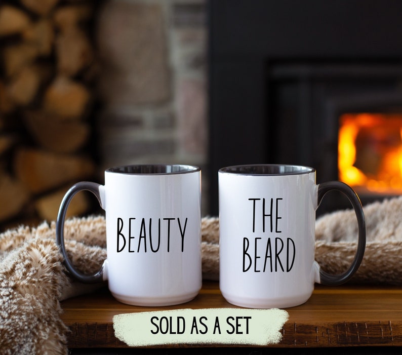 Two white coffee mugs with black handles and black rims. The mug on the left has the word 'BEAUTY' printed on it. The mug on the right has the words 'THE BEARD' printed on it. The mugs are on a wooden surface. The text 'SOLD AS A SET' is printed on a green banner below the mugs.