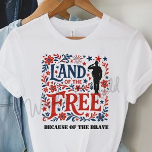 May include: White t-shirt with a patriotic design. The graphic features the words "LAND OF THE FREE" in blue and "FREE" in red, with a silhouette of a saluting soldier. The phrase "BECAUSE OF THE BRAVE" is below.
