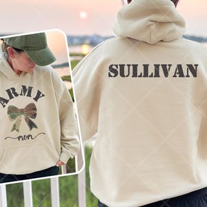 May include: A beige hoodie with the word "ARMY" arched above a camouflage bow graphic and the word "mom" below. The back of the hoodie has the name "SULLIVAN" printed across the shoulders. The person is wearing a green baseball cap.