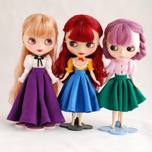 May include: Three dolls with different coloured hair and outfits. The doll on the left has blonde hair and is wearing a white blouse with a black bow, a purple skirt, and white shoes. The doll in the middle has red hair and is wearing a yellow blouse with a red bow, a blue skirt, and black shoes. The doll on the right has purple hair and is wearing a pink blouse with a white collar, a green skirt, and white shoes.