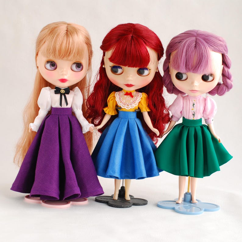 May include: Three dolls with different coloured hair and outfits. The doll on the left has blonde hair and is wearing a white blouse with a black bow, a purple skirt, and white shoes. The doll in the middle has red hair and is wearing a yellow blouse with a red bow, a blue skirt, and black shoes. The doll on the right has purple hair and is wearing a pink blouse with a white collar, a green skirt, and white shoes.