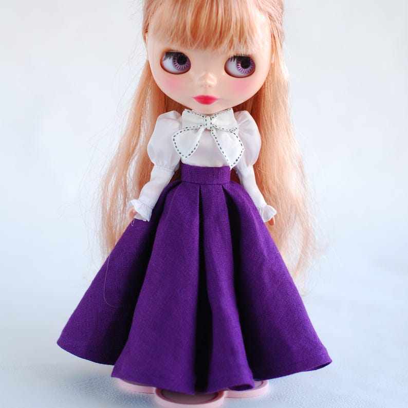 May include: A doll wearing a white blouse with a bow and a purple skirt.