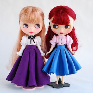 May include: Two dolls wearing dresses. The doll on the left has blonde hair and is wearing a white blouse with a black bow, a purple skirt, and white shoes. The doll on the right has red hair and is wearing a pink blouse with a floral pattern, a blue skirt, and black shoes.