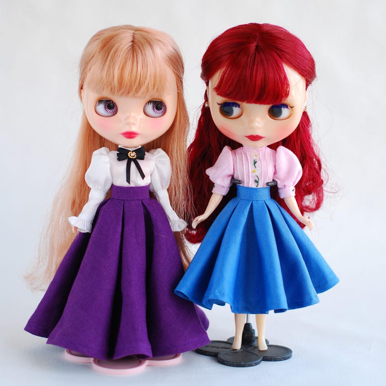May include: Two dolls wearing dresses. The doll on the left has blonde hair and is wearing a white blouse with a black bow, a purple skirt, and white shoes. The doll on the right has red hair and is wearing a pink blouse with a floral pattern, a blue skirt, and black shoes.